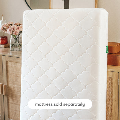The Newton Baby Extra Waterproof Crib Mattress Cover stands upright before a wooden dresser with flowers and decor. A label notes "mattress sold separately" and highlights the breathable protection of this white, quilted cover.