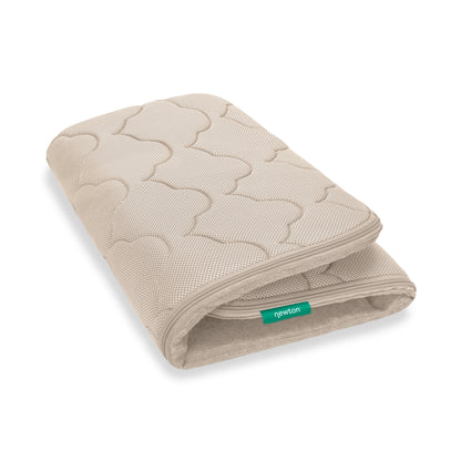 The Newton Baby Extra Bassinet Mattress Cover is beige and rectangular with a quilted wave pattern and a green "newton" tag. The machine-washable cover is partially folded for easy storage.
