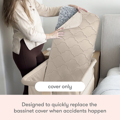 A person lifts a beige, quilted Newton Baby Extra Bassinet Mattress Cover to reveal its speckled underside beside a bed. Text reads: “cover only” and “Easily replace the Bassinet Mattress Cover after accidents. Machine washable.”.