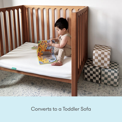 A young child sits in a Newton Galileo Mini Crib with Newton Mattress by Newton Baby, reading a picture book. Three soft, checkered cubes are stacked nearby. Text below reads: Converts to a Toddler Sofa.