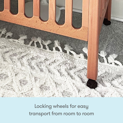 Close-up of a Newton Galileo Mini Crib with Newton Mattress leg by Newton Baby, featuring a black locking wheel on a white textured rug. Text below reads: “Locking wheels for easy transport from room to room.”.