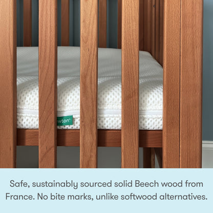A close-up of the Newton Galileo Mini Crib with Newton Mattress by Newton Baby showcases its solid Beech wood vertical slats and a white mattress with green Newton label, emphasizing superior safety and durability for a sturdy, portable crib.