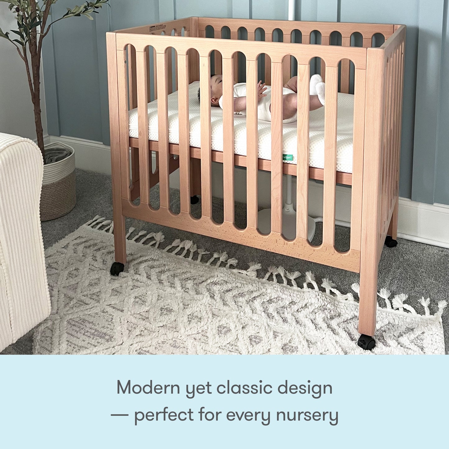 A baby rests in a Newton Galileo Mini Crib with Newton Mattress by Newton Baby, surrounded by a softly lit nursery featuring a patterned rug and cozy armchair. Text reads: “Modern yet classic design — perfect for every nursery.”.