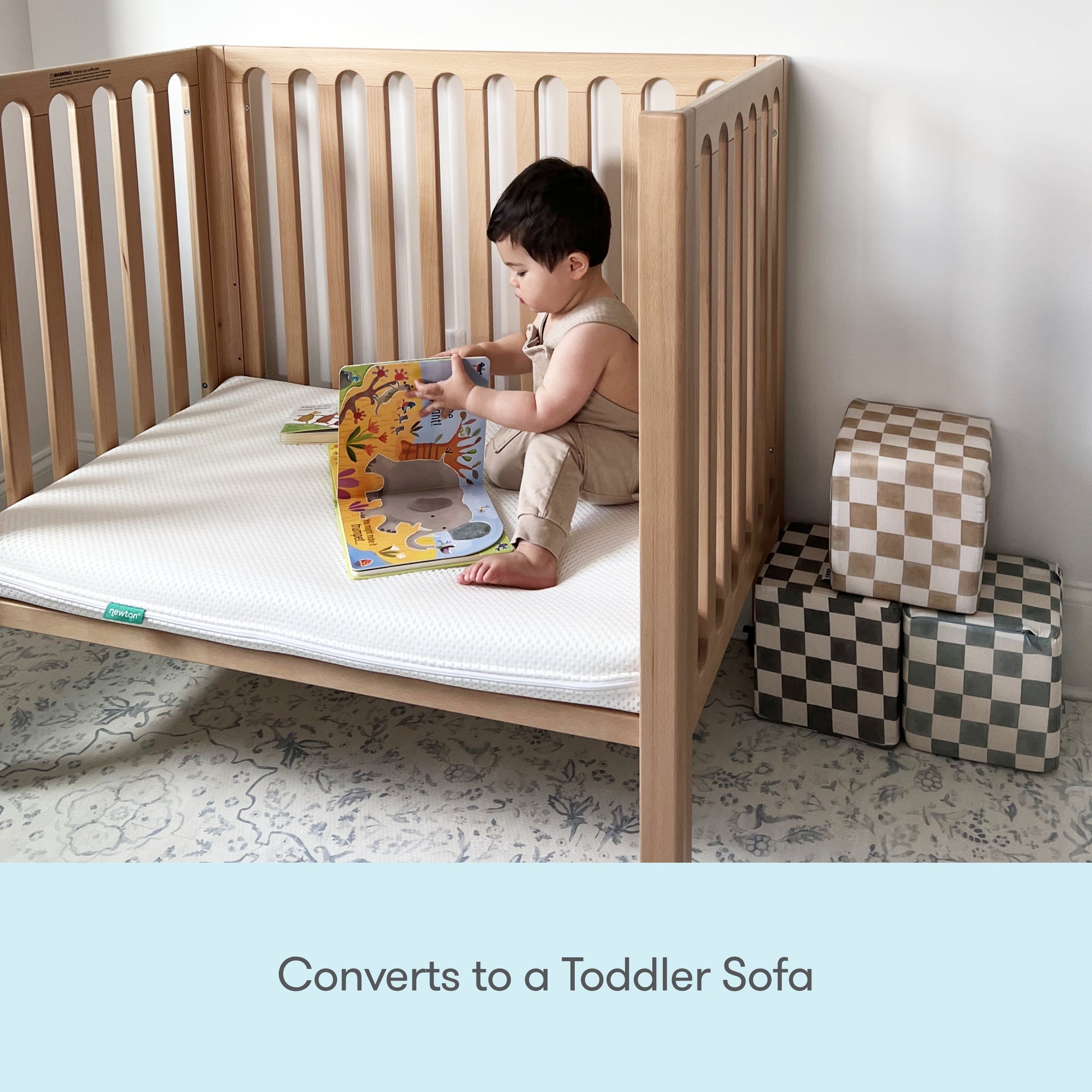 A young child sits in the Newton Galileo Mini Crib with Newton Mattress by Newton Baby, reading a picture book. Patterned soft blocks are stacked nearby. Text at the bottom reads: Converts to a Toddler Sofa.