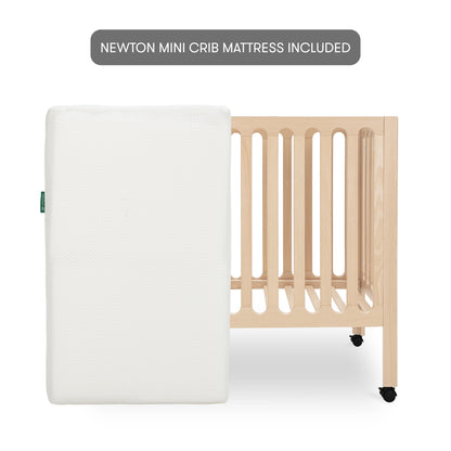 The Newton Galileo Mini Crib with Newton Mattress by Newton Baby, shown in light beech wood with wheels behind a white mattress, highlights the included NEWTON MINI CRIB MATTRESS and stylish, convenient portable design.