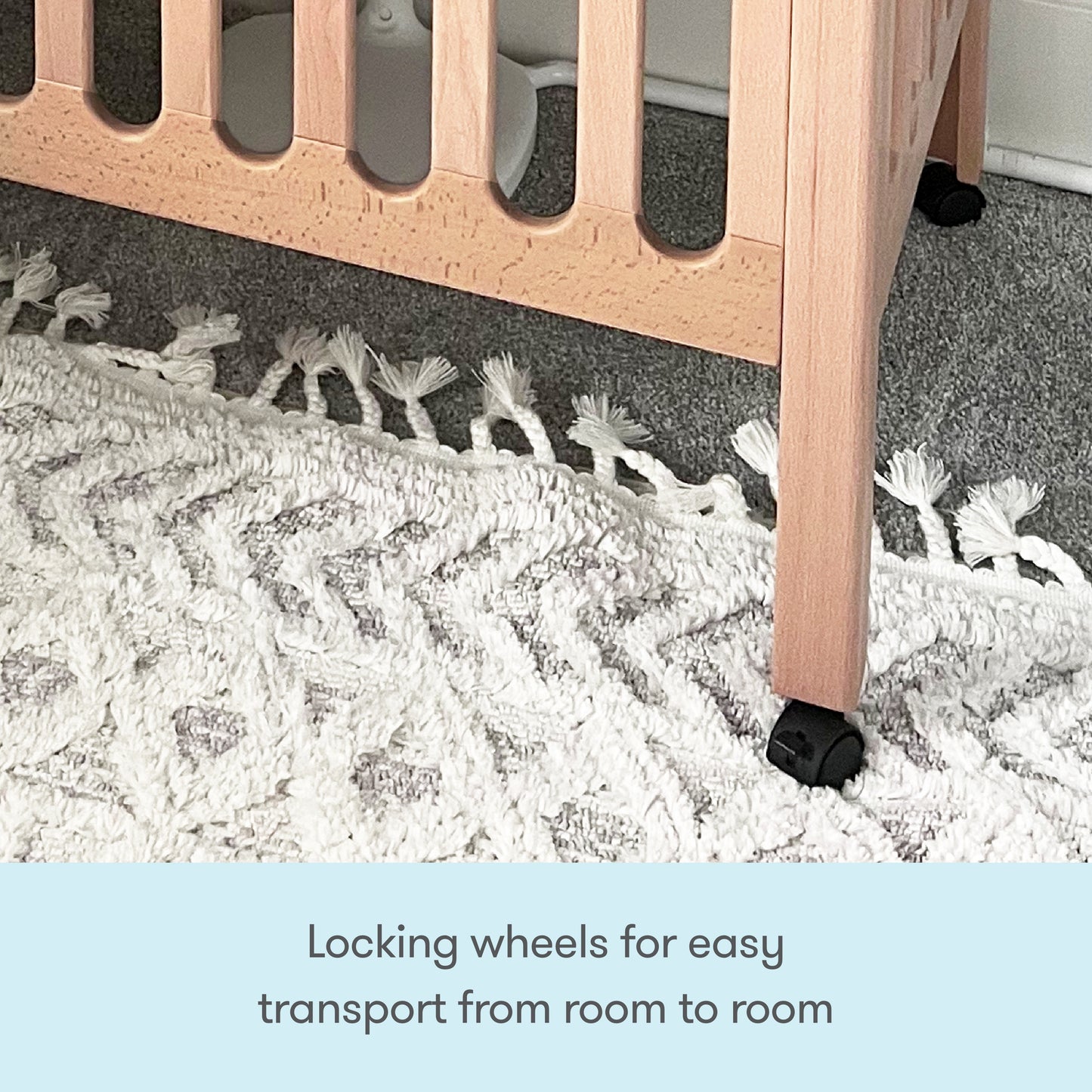 Close-up of a Newton Galileo Mini Crib with Newton Mattress by Newton Baby, showing its beech wood leg and black locking wheel on a white rug and gray carpet. Text below: Locking wheels make it easy to move from room to room.