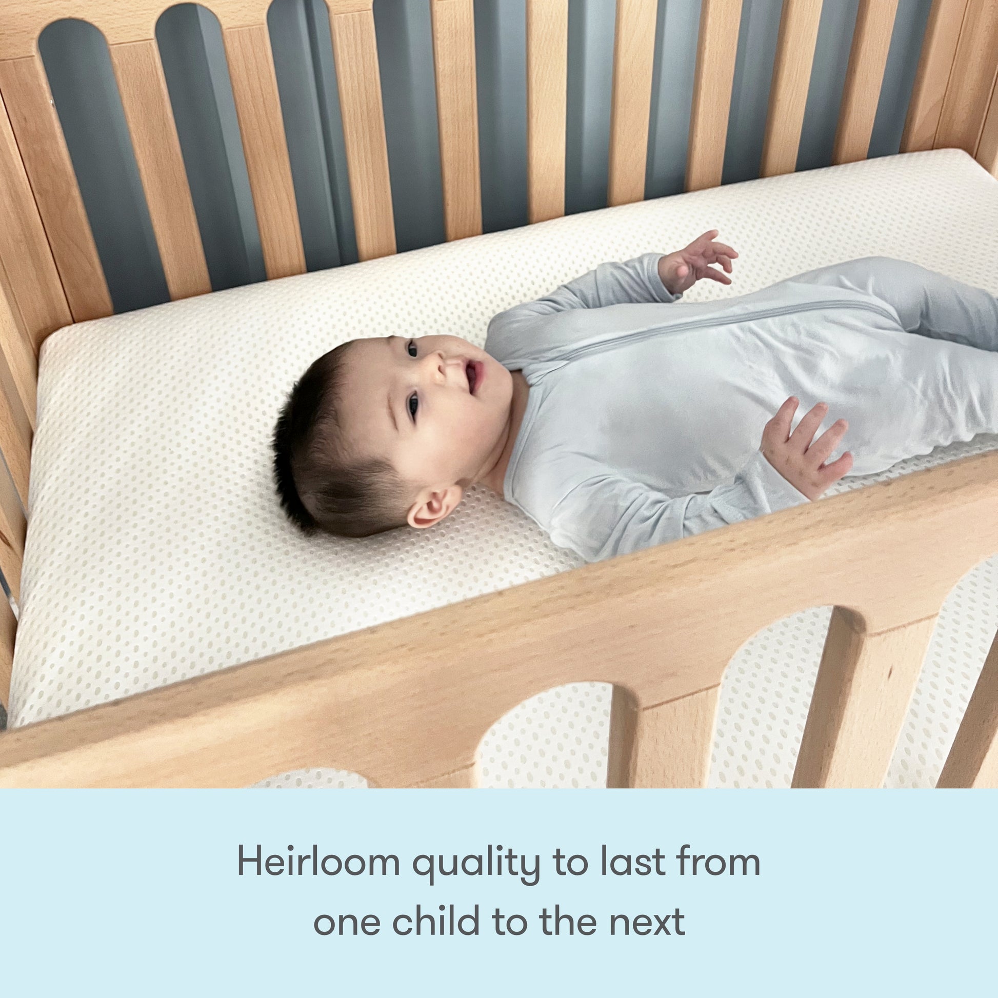 A baby in a light gray onesie lies on a Newton Galileo Mini Crib with Newton Mattress by Newton Baby, crafted from beech wood. Text below reads, Heirloom quality to last from one child to the next.
