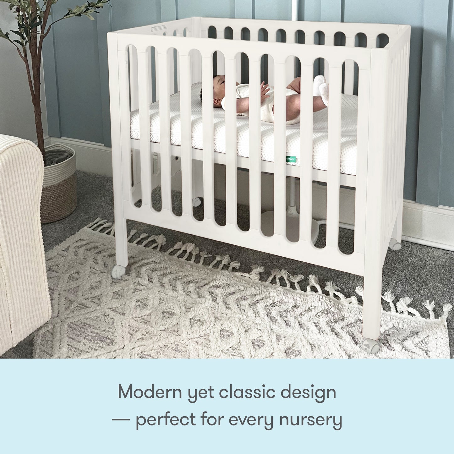 A baby rests in a Newton Galileo Mini Crib with Newton Mattress by Newton Baby, featuring vertical slats and set on a patterned rug against light blue paneled nursery walls. Text below reads: Modern yet classic design—perfect for every nursery.