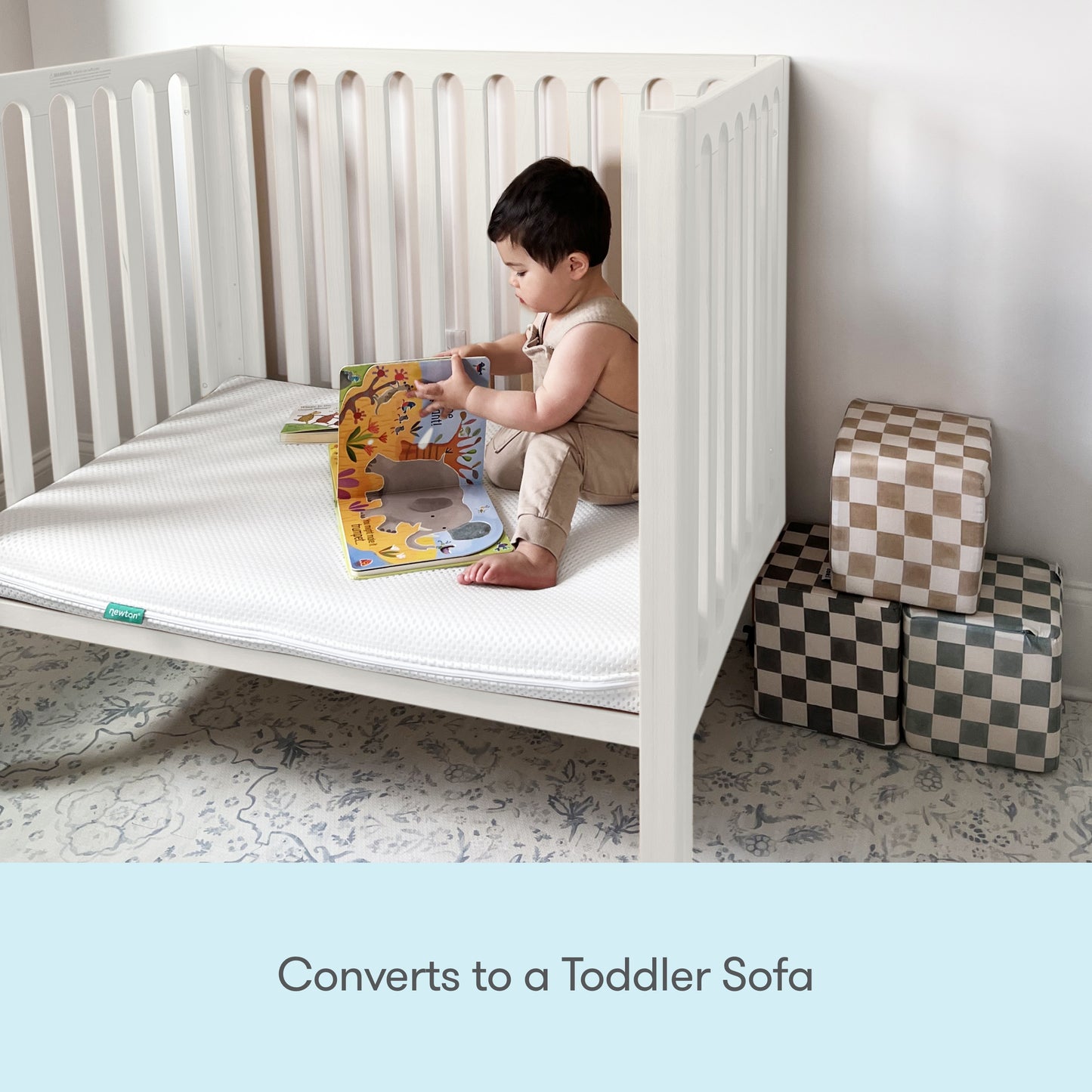 A young child sits inside a Newton Galileo Mini Crib with Newton Mattress by Newton Baby, reading a colorful picture book. Soft patterned cubes are stacked nearby. Text below reads, Converts to a Toddler Sofa.