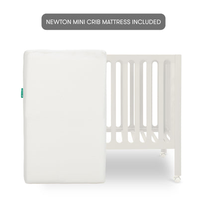 The Newton Galileo Mini Crib with Newton Mattress by Newton Baby is shown in white with vertical slats, and a white rectangular mattress leaning against it. A label above says, “NEWTON MINI CRIB MATTRESS INCLUDED.”.