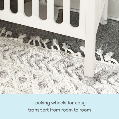 Close-up of the Newton Galileo Mini Crib with Newton Mattress by Newton Baby, showing its locking wheels on a textured white and gray rug. Text below: Locking wheels allow easy transport from room to room.
