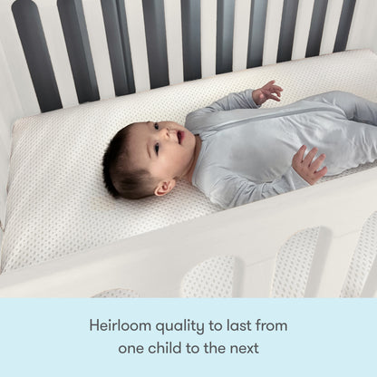 A baby in a light gray onesie lies on a white Newton Mattress inside the Newton Galileo Mini Crib by Newton Baby, featuring white rails with gray accents. Text below reads: Heirloom quality to last from one child to the next.