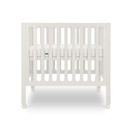 The Newton Baby Galileo Mini Crib with Newton Mattress is shown from the front, featuring white vertical slats, a mattress inside, and wheels for easy mobility against a plain white background.
