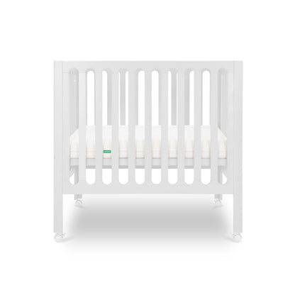 The Newton Baby Galileo Mini Crib with Newton Mattress features vertical slats, a comfy mattress, and four wheels. Crafted from durable beech wood, this portable white crib appears against a clean white background.