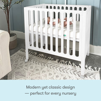 A baby rests in a white Newton Galileo Mini Crib with Newton Mattress by Newton Baby, featuring vertical slats and set on a patterned rug in a softly lit nursery. Modern yet classic design—perfect for every nursery.