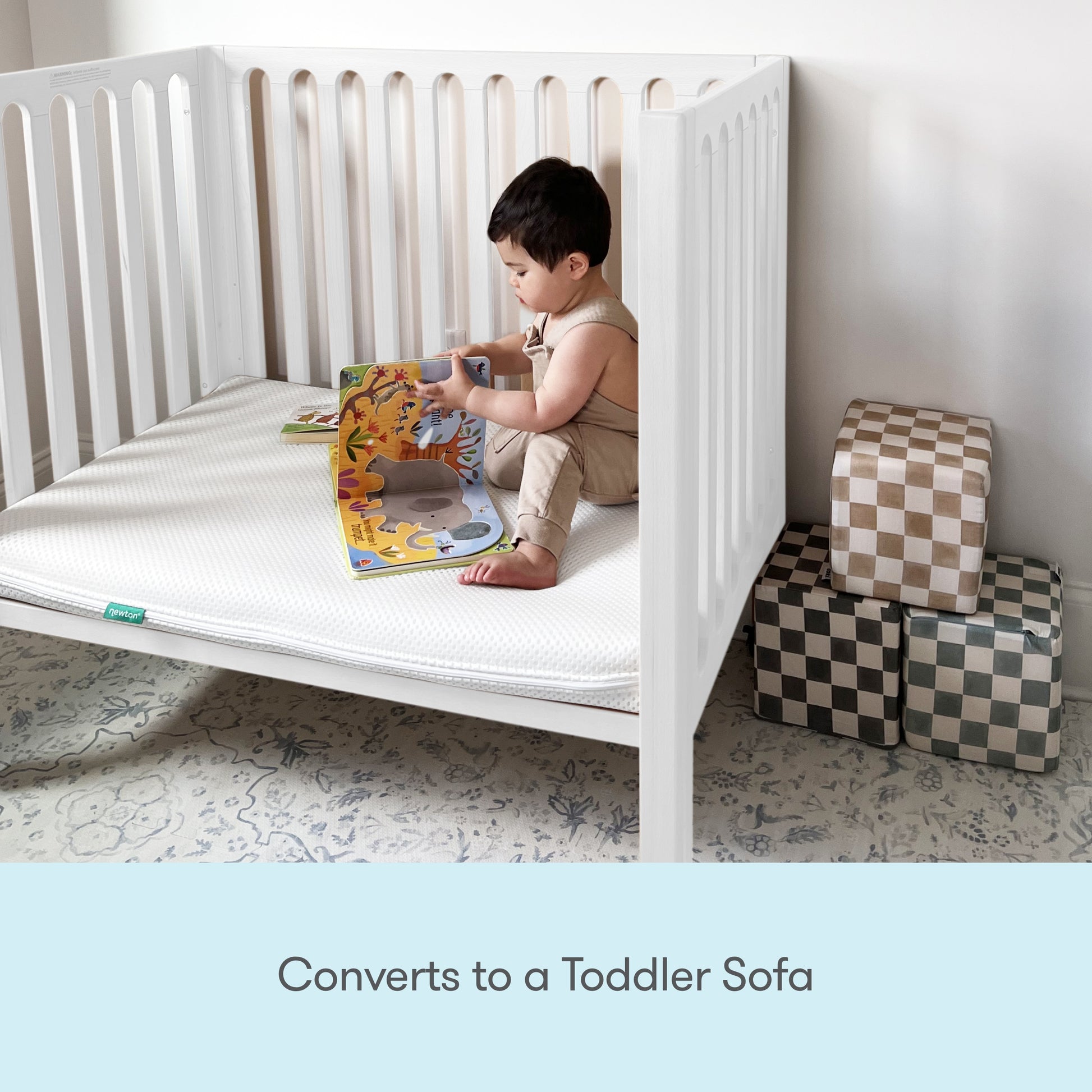 A young child sits in a white Newton Galileo Mini Crib with Newton Mattress by Newton Baby, reading a colorful book. Two checkered soft blocks are beside the crib. Text below reads, Converts to a Toddler Sofa.