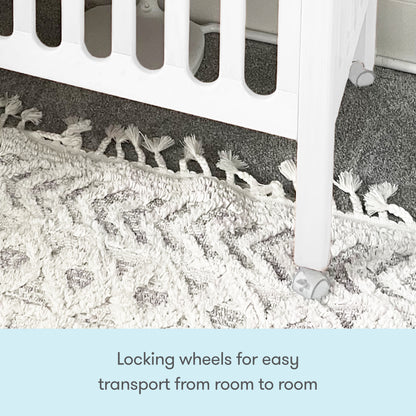 A close-up shows the bottom corner of the Newton Galileo Mini Crib with Newton Mattress by Newton Baby on locking wheels, set on a white rug and grey carpet. Text reads: Locking wheels make it easy to move between rooms.