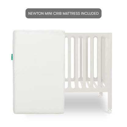 Warm White modern baby crib on wheels with a mattress stood up in front of it with text "newton mini crib mattress included".