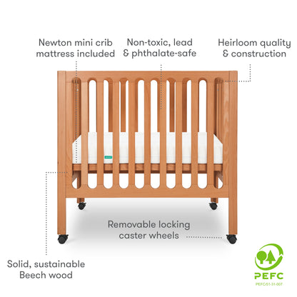Modern wooden crib on wheels with visible mattress, isolated on a white background. With text "newton mini crib mattress included, non-toxic, lead & phthalate-safe, heirloom quality & construction, removable locking caster wheels, solid, sustainable beech wood." PEFC Certified