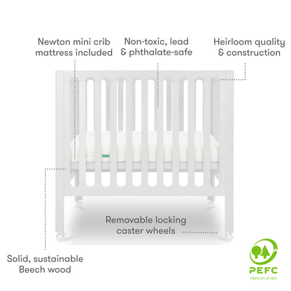 White modern crib on wheels with visible mattress, isolated on a plain background. With text "newton mini crib mattress included, non-toxic, lead & phthalate-safe, heirloom quality & construction, solid sustainable beech wood, removable locking caster wheels, PEFC Certified"