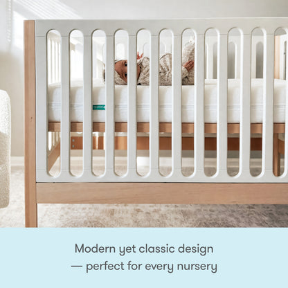 A baby rests in the Newton Galileo Convertible Crib by Newton Baby, featuring beech wood and white slats in a softly lit nursery. Text: Modern yet classic—this crib is the perfect addition to any nursery.