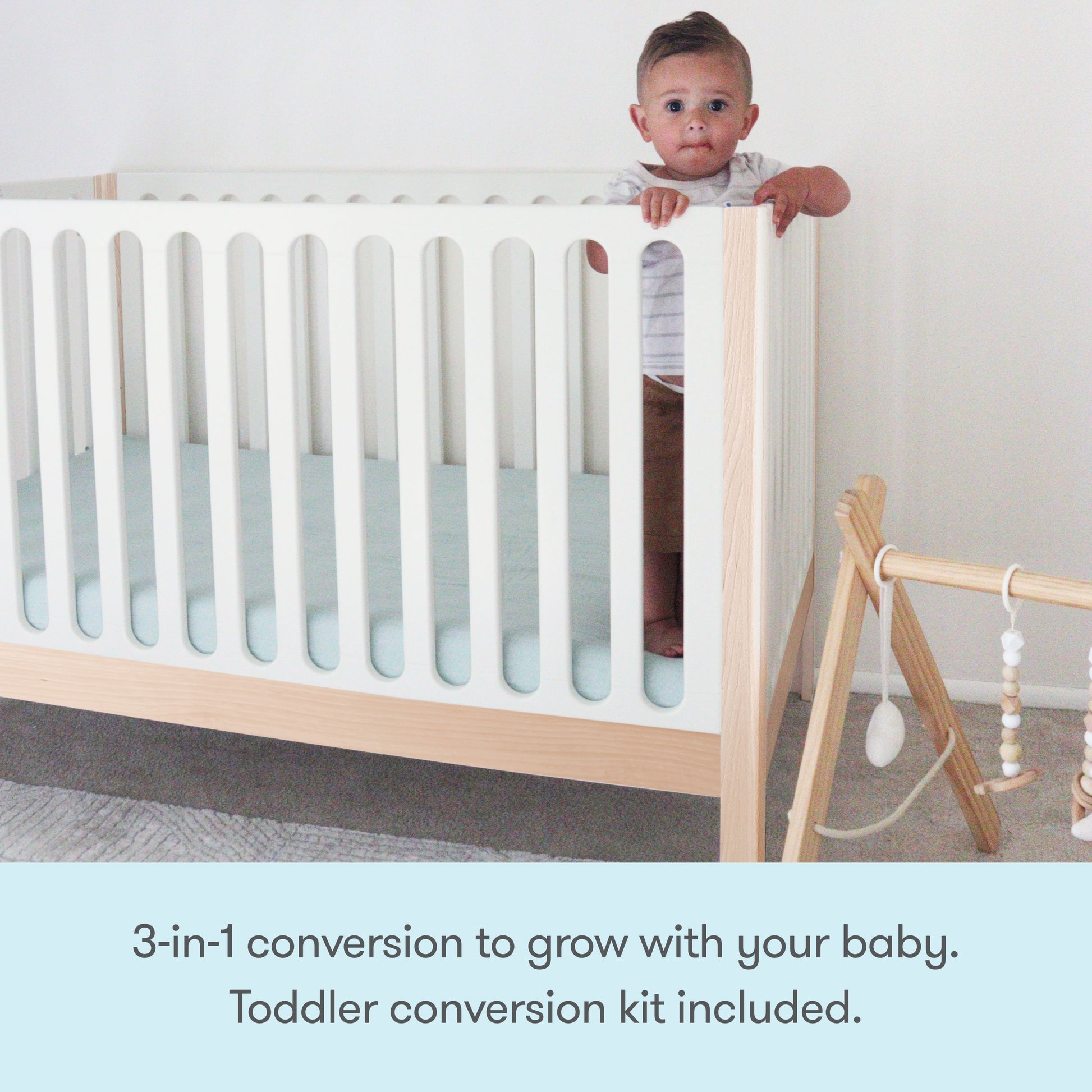 A baby stands holding the Newton Galileo Convertible Crib by Newton Baby in a minimalist nursery. Nearby is a wooden baby gym. Text reads: 3-in-1 convertible design grows with your child—toddler bed conversion kit included.