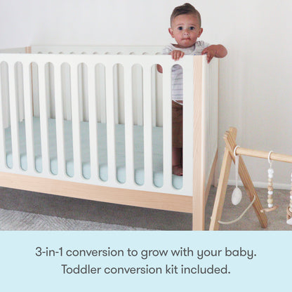 A baby stands holding the Newton Galileo Convertible Crib by Newton Baby in a minimalist nursery. Nearby is a wooden baby gym. Text reads: 3-in-1 convertible design grows with your child—toddler bed conversion kit included.