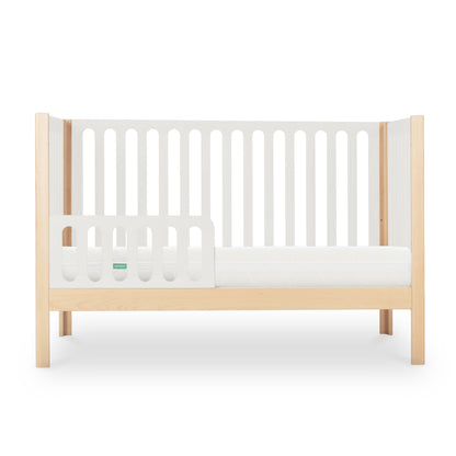 The Newton Galileo Convertible Crib by Newton Baby features light beech wood legs, white slatted sides, and a partial side rail. This stylish 3-in-1 crib easily converts to a toddler bed. Shown with a white mattress against a plain background.