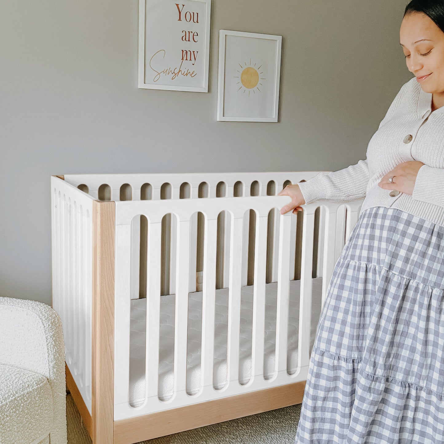 A pregnant woman stands beside the Newton Galileo Convertible Crib by Newton Baby in a softly lit nursery with framed wall art and neutral decor, highlighting modern nursery furniture. 999-8402-NDW