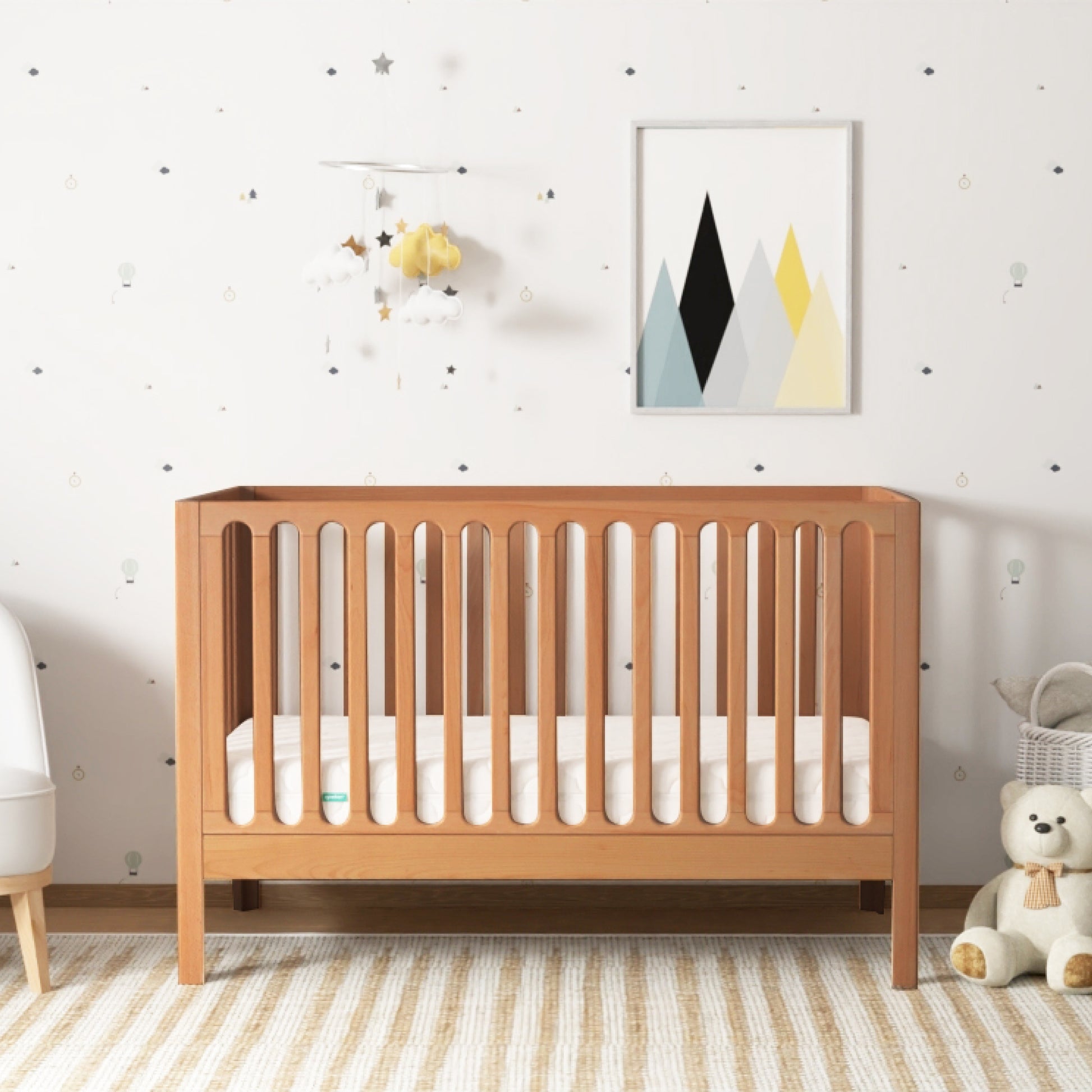 The Newton Galileo Convertible Crib by Newton Baby with a white mattress sits against patterned wallpaper. A geometric mountain art print and mobile hang above, while a teddy bear rests on the striped rug beside the crib. 999-8420-CST