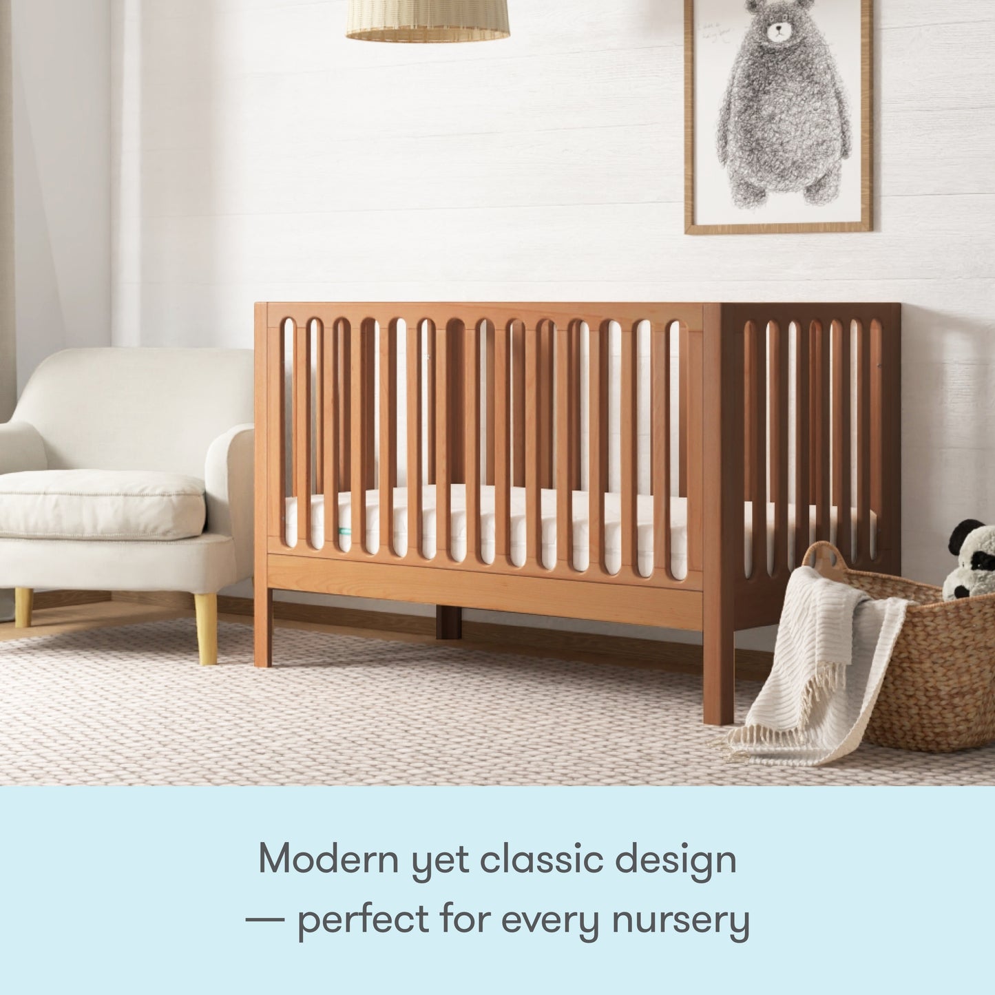 The Newton Galileo Convertible Crib by Newton Baby, made of beech wood with vertical slats, stands beside a white sofa and woven basket in a cozy nursery. A framed bear print hangs above. Modern yet classic—perfect for every nursery.