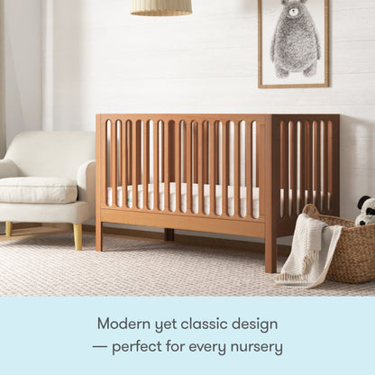 The Newton Galileo Convertible Crib by Newton Baby, made of beech wood with vertical slats, stands beside a white sofa and woven basket in a cozy nursery. A framed bear print hangs above. Modern yet classic—perfect for every nursery.