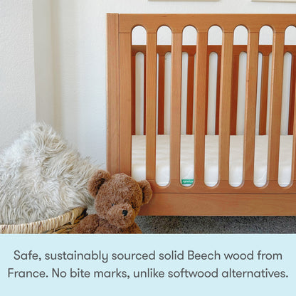 The Newton Galileo Convertible Crib by Newton Baby, made from solid beech wood, offers enhanced safety, durability, and easy toddler bed conversion compared to softwood cribs. A basket with a fuzzy blanket and brown plush bear sits beside it.