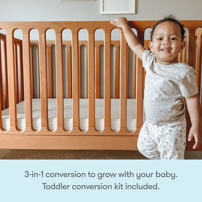 A smiling toddler stands next to the Newton Galileo Convertible Crib by Newton Baby. Text below reads: 3-in-1 crib grows with your child—toddler bed conversion kit included.