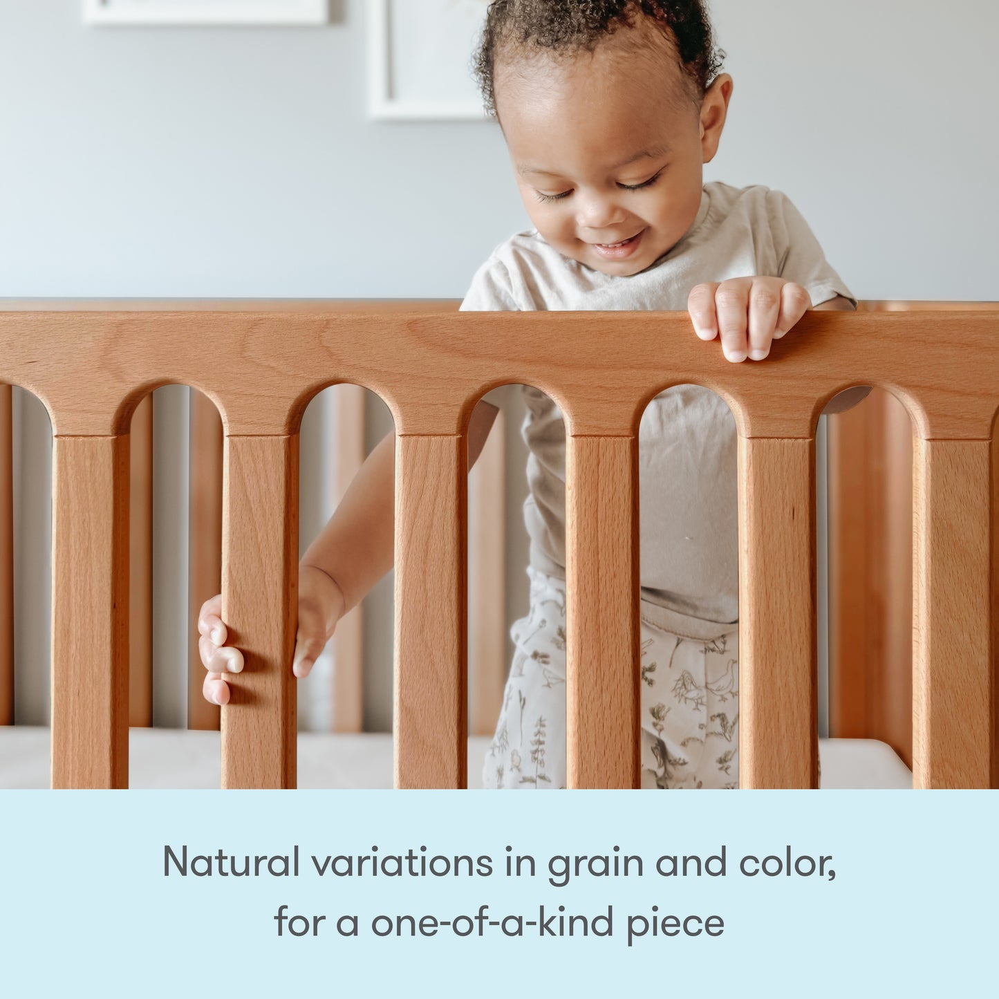 A happy toddler stands in a Newton Galileo Convertible Crib by Newton Baby, gripping the top rail. Text below reads: “Unique wood grain and color make each crib one-of-a-kind.”.