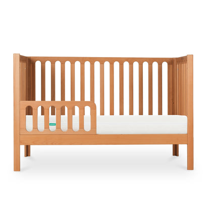 The Newton Galileo Convertible Crib by Newton Baby is a beech wood crib with vertical slats, featuring a partial guardrail and white mattress. Shown in toddler bed mode, it’s photographed against a white background.