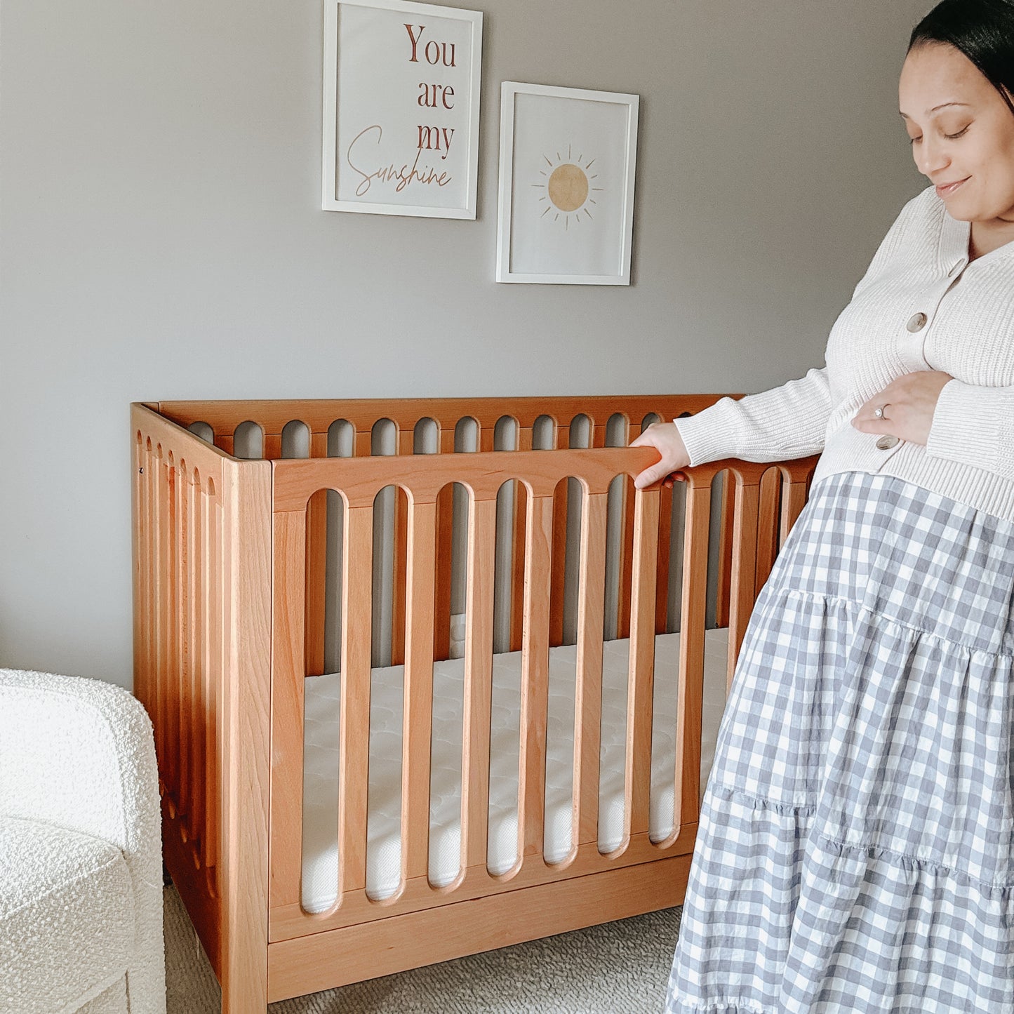 A woman stands beside a Newton Galileo Convertible Crib by Newton Baby in a softly lit nursery, gently touching her belly. Two framed prints hang above—one says “You are my sunshine,” the other features a simple sun illustration.