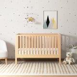 In the nursery, the Newton Baby Newton Galileo Convertible Crib stands beneath playful wallpaper and geometric art, with a patterned rug, white armchair, basket, and teddy bear nearby—a stylish centerpiece of comfort and versatility. 999-8420-NAT