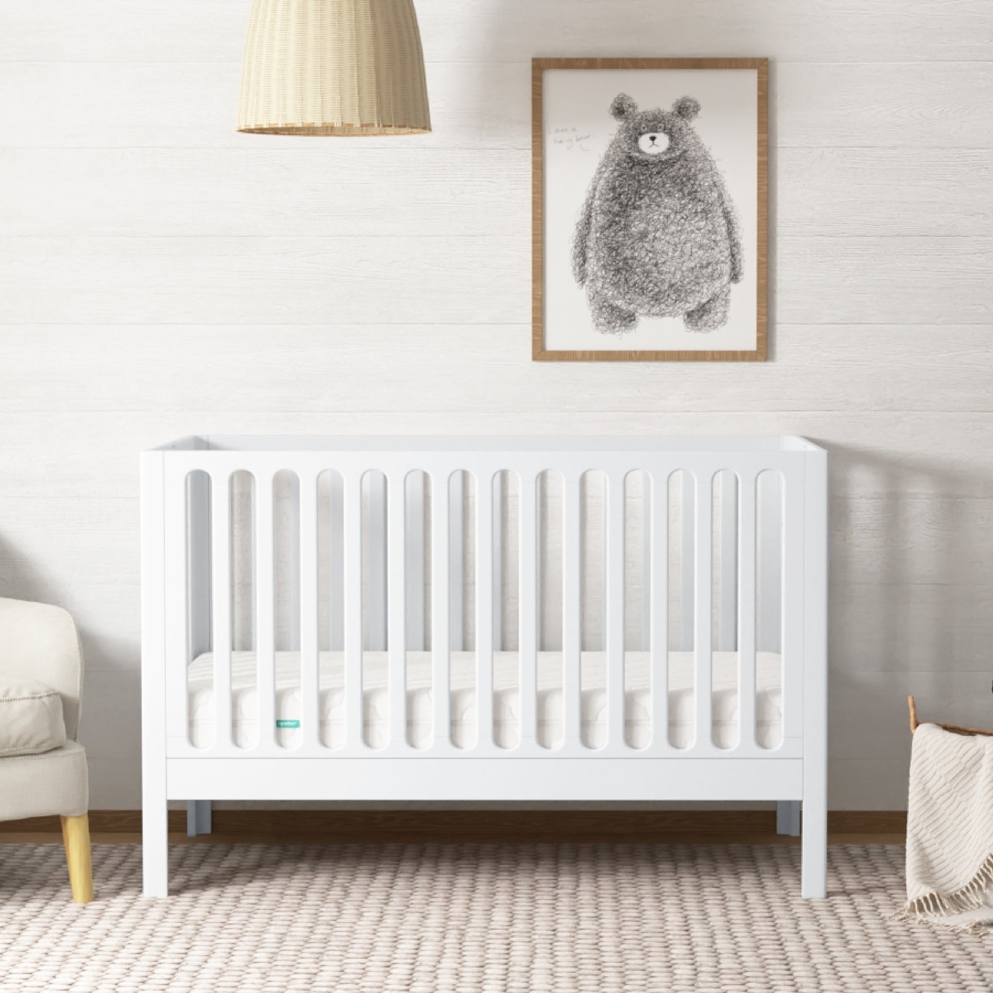 The Newton Galileo Convertible Crib by Newton Baby, made of white beech wood with vertical slats, sits in a softly lit nursery. A woven pendant lamp hangs above the crib, which converts to a toddler bed, surrounded by cozy décor and a bear print. 999-8420-WHT