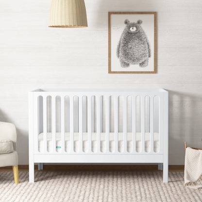 The Newton Galileo Convertible Crib by Newton Baby, made of white beech wood with vertical slats, sits in a softly lit nursery. A woven pendant lamp hangs above the crib, which converts to a toddler bed, surrounded by cozy décor and a bear print. 999-8420-WHT