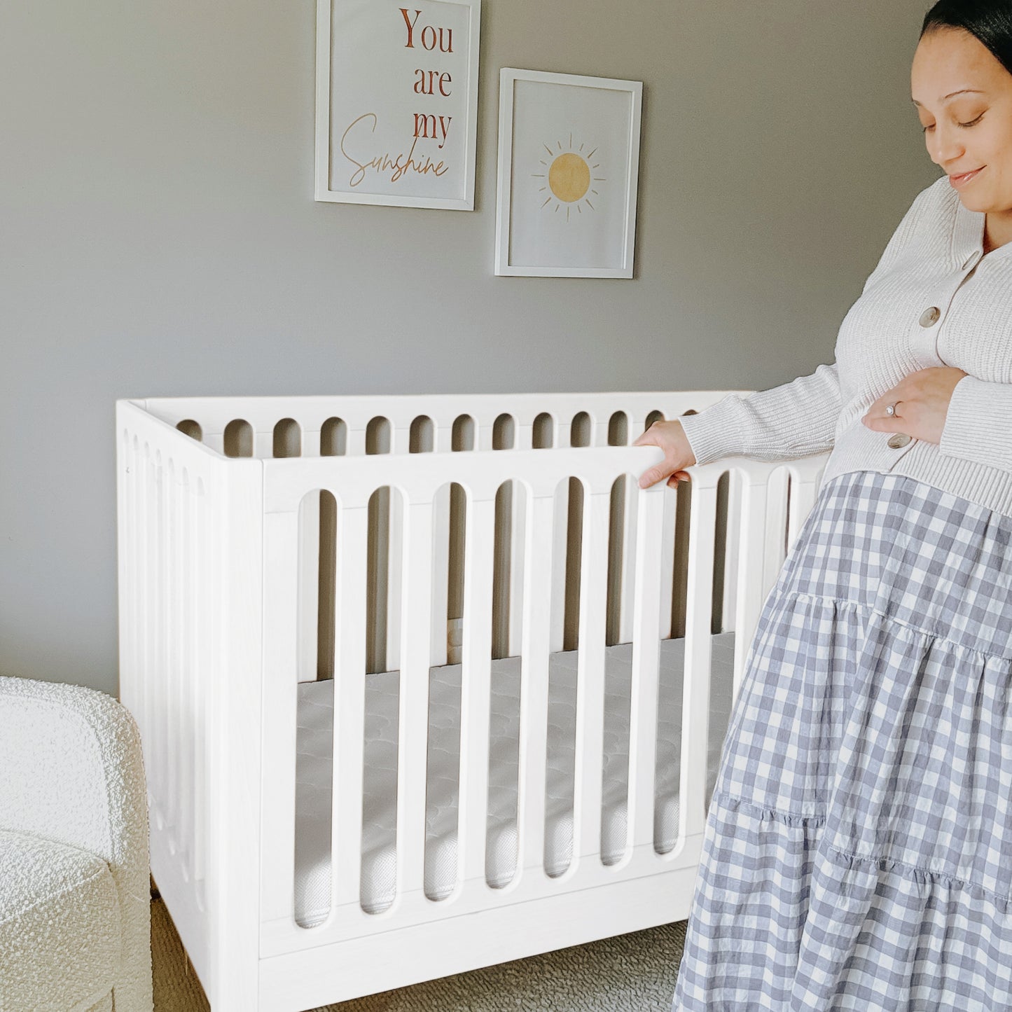 A pregnant woman stands beside the Newton Galileo Convertible Crib by Newton Baby in a softly lit nursery. Above the crib, two framed prints are hung: one features a sun illustration, the other reads “You are my Sunshine.”.