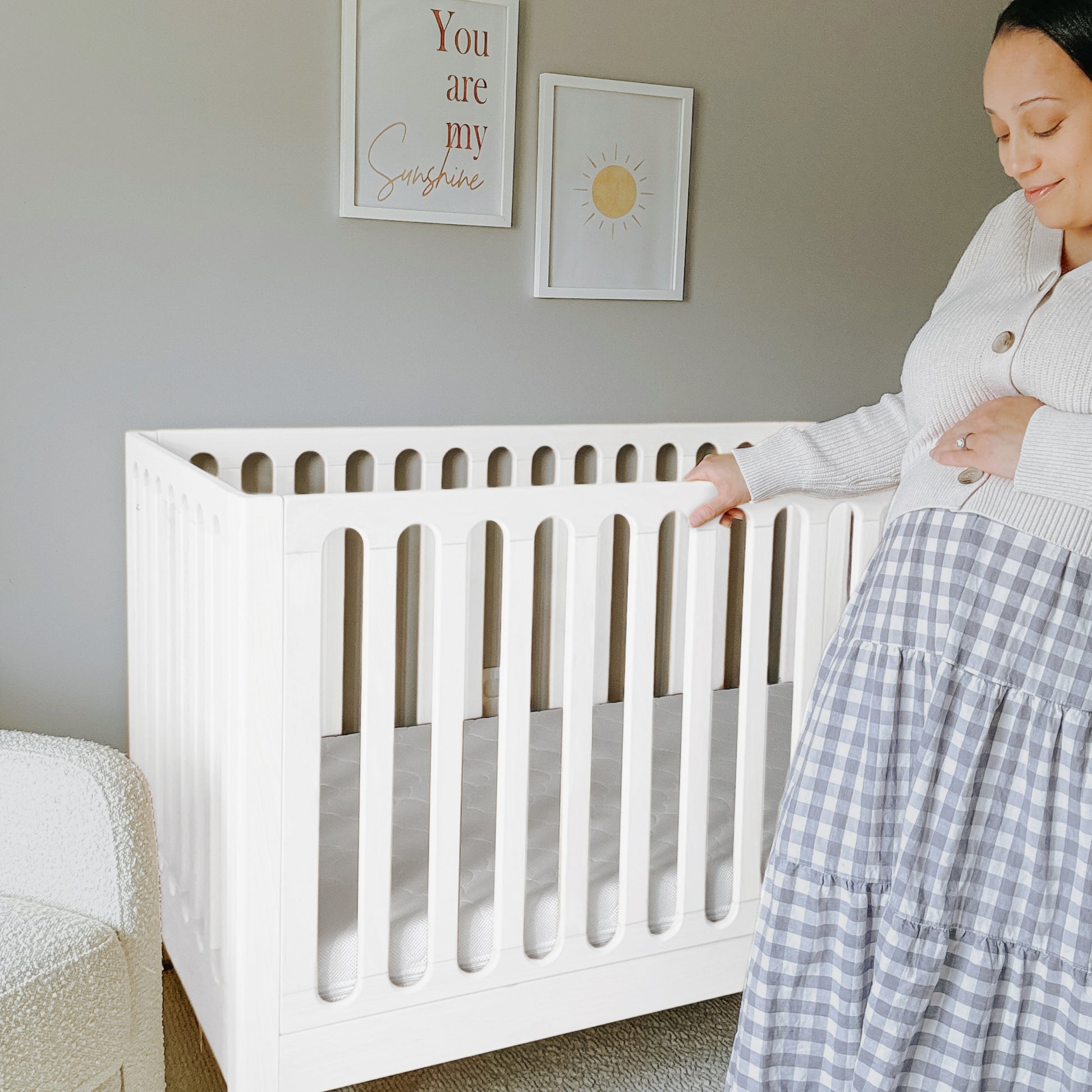 A pregnant woman stands beside the Newton Galileo Convertible Crib by Newton Baby in a softly lit nursery. Above the crib, two framed prints are hung: one features a sun illustration, the other reads “You are my Sunshine.”.