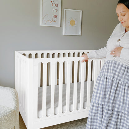 A pregnant woman stands beside the Newton Galileo Convertible Crib by Newton Baby in a softly lit nursery. Above the crib, two framed prints are hung: one features a sun illustration, the other reads “You are my Sunshine.”.