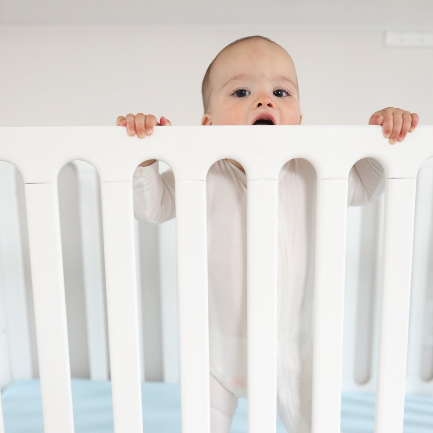 A baby in a white onesie stands inside the Newton Galileo Convertible Crib by Newton Baby, holding the rail with both hands and looking forward with their mouth slightly open.