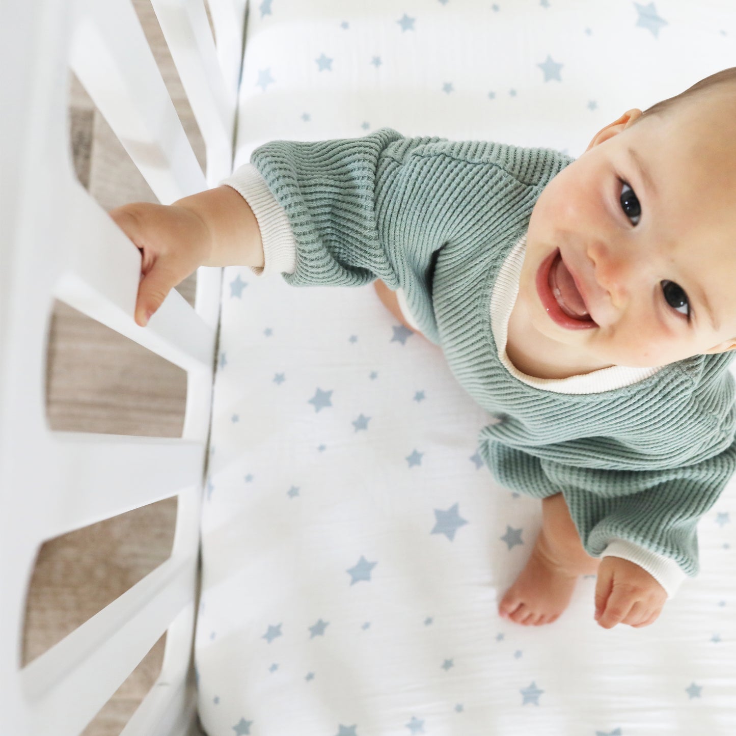 A smiling baby in a green sweater stands in a Newton Baby Newton Galileo Convertible Crib, holding the white railing. The white crib sheet has blue stars. Photo is taken from above.