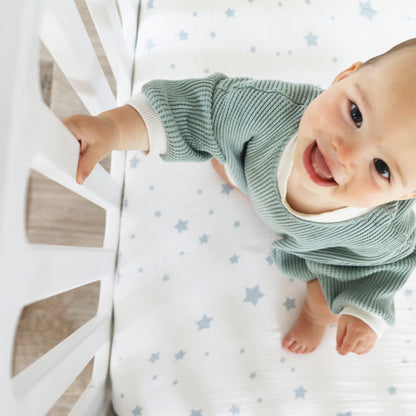 A smiling baby in a green sweater stands in a Newton Baby Newton Galileo Convertible Crib, holding the white railing. The white crib sheet has blue stars. Photo is taken from above.