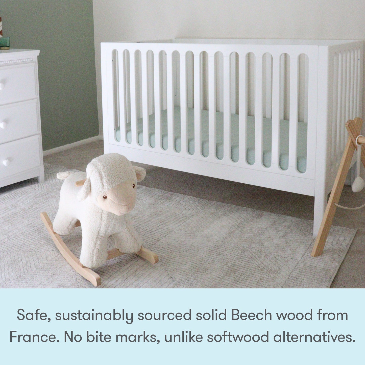 A white Newton Galileo Convertible Crib by Newton Baby on a light rug next to a dresser, with a plush rocking sheep in front. Caption: “Crafted from safe, sustainably sourced solid French Beech wood—no bite marks like softwood cribs.”.