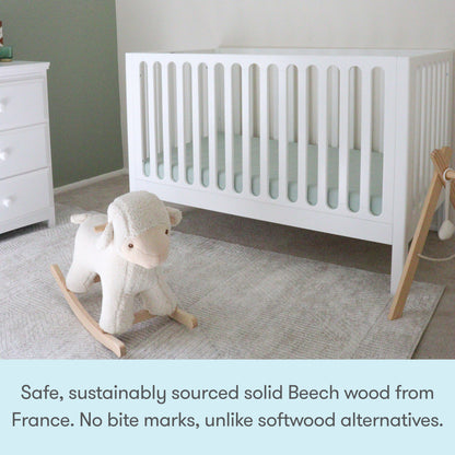 A white Newton Galileo Convertible Crib by Newton Baby on a light rug next to a dresser, with a plush rocking sheep in front. Caption: “Crafted from safe, sustainably sourced solid French Beech wood—no bite marks like softwood cribs.”.