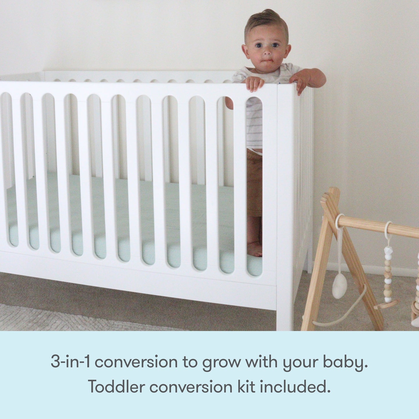 A baby stands by the Newton Galileo Convertible Crib by Newton Baby in a minimal nursery. Nearby is a wooden play gym. Text: “3-in-1 convertible crib grows with your baby. Toddler bed conversion kit included.”.