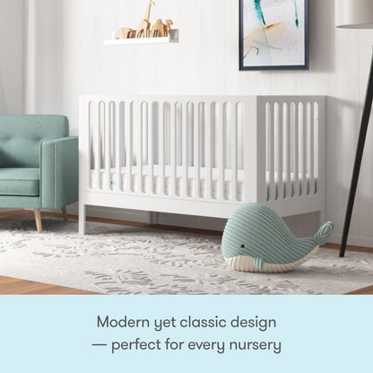 A stylish nursery featuring the Newton Galileo Convertible Crib by Newton Baby in white beech wood, a green armchair, plush whale toy, and soft rug. Modern decor and framed art complete the cozy, inviting space—modern yet classic for any nursery.
